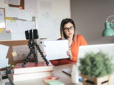 A woman teaches an online class. She holds a clipboard up facing her laptop, which is open on the desk in front of her. There are papers on the wall behind her and a tripod smartphone stand on her desk with books.