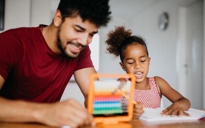非洲民族家庭在家里的客厅一起做作业。父亲帮助女儿做学前作业。拿算盘的男人和女孩正在笔记本上写字。