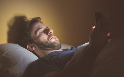 有面部头发的一个白色人在看他的巧妙的电话屏幕的床上。