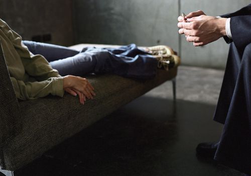 Young woman lying on couch and psychotherapist leaning toward her with hands clasped