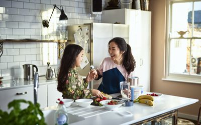 妇女在匙子拿着酸奶夫人往女孩，笑，威胁在女儿的面孔的污迹食物，混乱，获得乐趣，玩，粘合，粘合