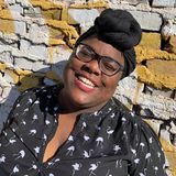 Taneasha standing front of white and yellow brick wall, wearing black headwrap and smiling