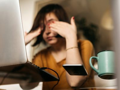 A young white person with chin-length brown hair sits at her laptop and rubs her eyes.