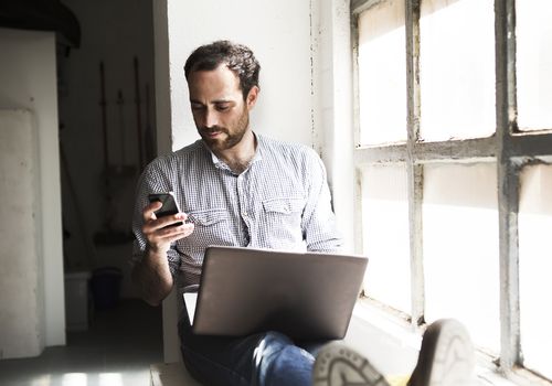 A man looking at his phone and computer, sitting on a windowsill