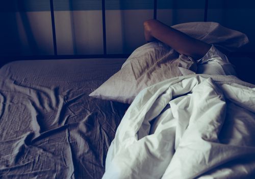Person in bed covered in sheets with a pillow over their head