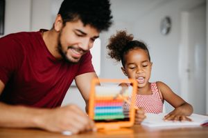 非洲裔家庭在客厅里一起做家庭作业。父亲帮助女儿做学龄前的家庭作业。男人拿着算盘，女孩在笔记本上写字。