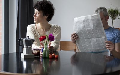 一个严肃的女人看着窗外，而她的男朋友躲在报纸后面