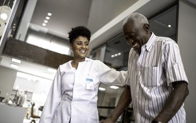 穿着白色外套的看护人有助于一个人使用助行器。两个人都在微笑。