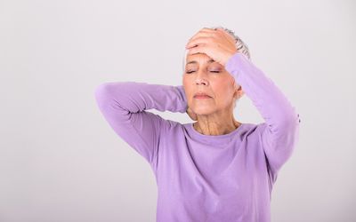 一名戴着紫色衬衫的资深妇女用双手握住她的头，闭上她的眼睛不适。