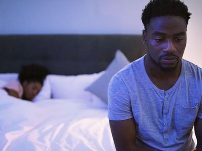 Sad man sits at edge of the bed while woman partner sleeps