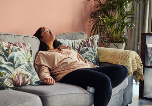 A stress-out woman plops down on her gray couch with her arms outstretched, looking up toward the ceiling.