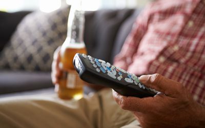 男人坐在沙发上拿着电视遥控器和一瓶