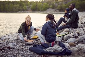 Mother preparing food on barbecue grill amidst family sitting on rocks at beach during camping