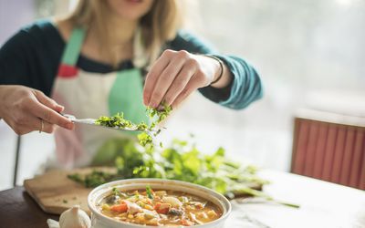 A woman wearing an apron, sitting at a table, sprinkling herbs into a bowl of vegetable stew.