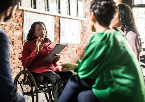 A black person using a wheelchair gives a presentation to colleagues at work.