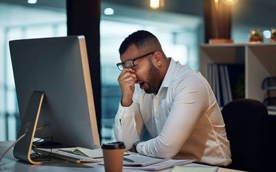 一个穿着白衬衫的男人坐在办公桌前，打着哈欠，揉着眼睛