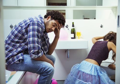 Man sitting on the edge of a bathtub while a woman vomits in a toilet