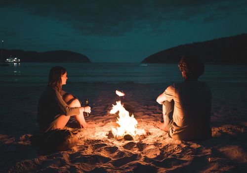 man and woman sitting by a campfire on a beach at night