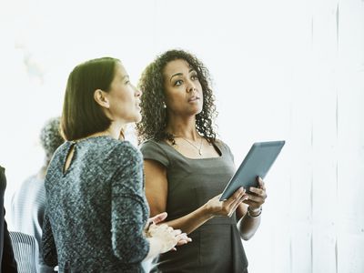 Two businesswomen look up at screen. One is holding an iPad.