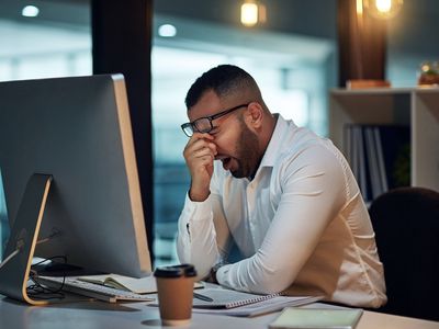 Man in white shirt sitting at his desk, yawning and rubbing his eyes